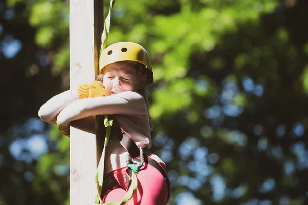 Um parque de cordas. Menina feliz escalando uma árvore. Rede de carga de escalada e pendurado log. Caminho da corda - menina está olhando para o slideway corda temendo . — Fotografia de Stock