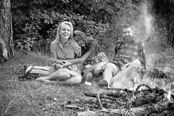 Bearded man and women smile at bonfire. Happy friends at campfire. Hipster in palid shirt and girls in vintage dresses relax in forest. Summer vacation concept. Camping travel and wanderlust, retro