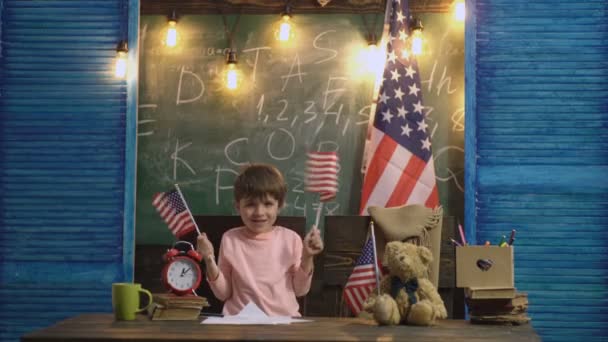 Garçon en classe agitant des drapeaux américains sur le fond de la commission scolaire et grand drapeau des États-Unis. Joyeux petit garçon agitant des drapeaux américains sur fond de classe. Naissance du patriotisme chez les jeunes enfants . — Video