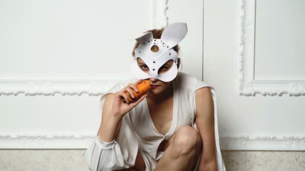 A girl dressed as a rabbit licks and nibble carrots isolated on white. Funny brutal bunny girl. Nibbles a carrot like a hare. Ready for the party. Pretty, trendy woman is wearing bunnies leather mask.