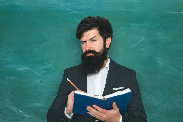 Estudiante masculino serio estudiando en la escuela. Joven profesor barbudo cerca de pizarra en el aula de la escuela. Profesor barbudo en clase de la escuela en los escritorios en el aula . —  Fotos de Stock