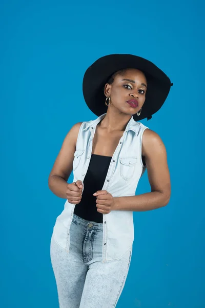 Lady on calm face with makeup. Lady in modern clothes as denim vest and jeans. Fashion concept. Woman with african appearance in black hat and denim clothes looks gorgeous and stylish blue background