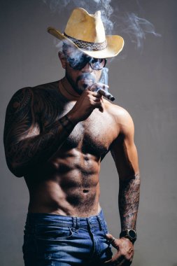 Sağlık. Sigara içiyor. duman üfler. kötü alışkanlık. güven karizması. dövme adam seksi abs. erkek modası. atletik vücutile kaslı maço adam. kovboy şapkalı acımasız sporcu.