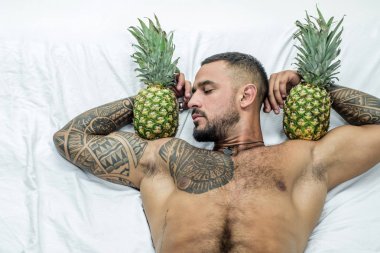Kaslı adam ananas meyvesini pazıların üzerinde tutuyor. Vahşi sporcu yatakta rahatla. Steroitler. Seksi dövmeli adam diyeti. Erkek sağlığı. Atletik vücudu olan kaslı maço bir adam. egzotik meyve sağlıklı yiyecek vitamini.
