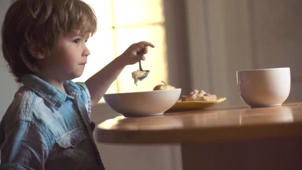 Boldog baba fiú kanál megeszi magát. Kisfiú ül az asztalnál, és étkezési tej snack. Baby étkezési élelmiszer a konyhában. Gyermekgondozási. Kisfiú ül az asztalnál, és étkezési tej snack. — Stock videók