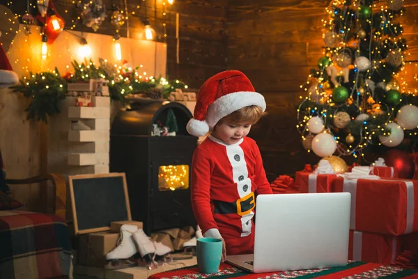 O miúdo está a usar roupa de Pai Natal junto ao portátil. Conceito de Natal. Fundo da lareira. Luz de Natal . — Fotografia de Stock