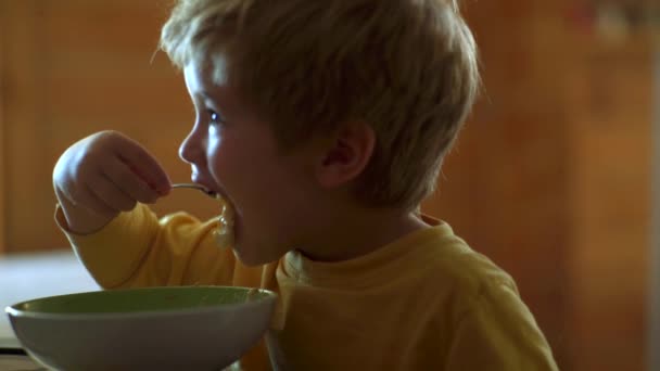 食卓の台所にいる子供。幸せな子供だ陽気な赤ちゃんの子供はスプーンで食べ物自体を食べる. — ストック動画