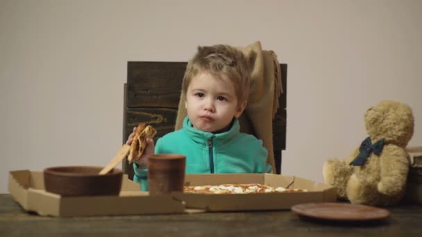 Lindo niño caucásico comiendo pizza en la mesa de madera en la que hay caja de pizza, cuchara, plato y oso aislado en blanco. Niño hambriento tomando un bocado de pizza. Servicio de reparto de comida. Antecedentes . — Vídeo de stock