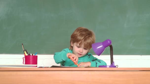 Um rapazinho engraçado a apontar para o quadro negro. Miúdos da escola. Criança sorridente alegre no quadro negro. Educação. Crianças na aula da escola. Sala de aula. Alunos sorridentes felizes desenhando na mesa . — Vídeo de Stock