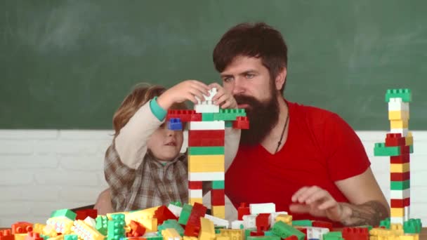 Toddler boy in an orange helmet or helmet helps his bearded father. Little kid and his father building House Together. Child with her parent in Hard Hat Playing Building Blocks. — Stock Video