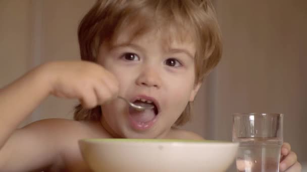 Mały chłopiec, który ma śniadanie w kuchni. Małe dziecko jedzą. Głodny mały chłopiec jedzenia. Młody dzieciak siedzi na stole jedzenia zdrowej żywności z zabawnym wyrazem na twarzy. — Wideo stockowe