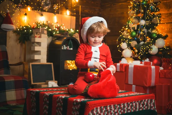 クリスマスストッキング。クリスマスの奇跡と新年の気持ち。暖かい服を着た小さな男の子が座って、織物のおもちゃで遊んでいます。オープンエンドのゲーム。子供の頃の最初の思い出. — ストック写真