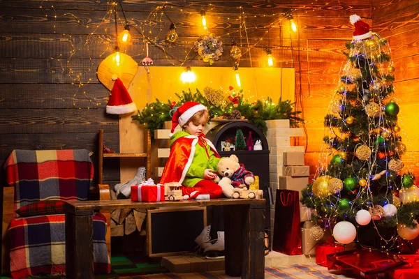 Niño sentado en una mesa de madera y jugando con regalos. Juguetes cerrados y abiertos. Juguetes naturales de madera y textiles para niños juegan. Árbol de Navidad y decoraciones de año nuevo . —  Fotos de Stock