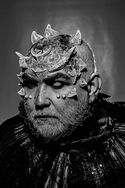 Demon with golden collar on black background. Man with thorns or warts, face covered with glitters. Fantasy concept. Alien, demon, sorcerer makeup. Senior man with white beard dressed like monster.