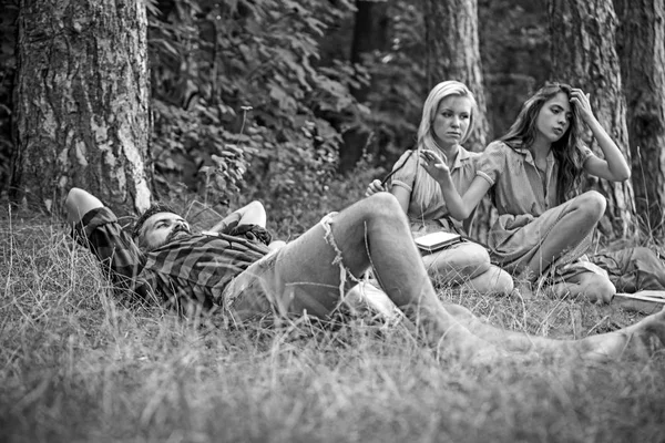 Ισχυρό άντρα ξαπλωμένος στο γρασίδι. Δύο κορίτσια διαβάζοντας βιβλία δίπλα από την πυρά προσκόπων. Φίλοι κάμπινγκ το καλοκαίρι — Φωτογραφία Αρχείου