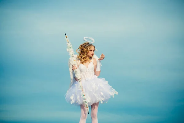 A beautiful teen with blonde curly hair and a bow and arrow as cupid - Valentines Day. Little angel in love. Space for text. Portrait of a cupid little girl