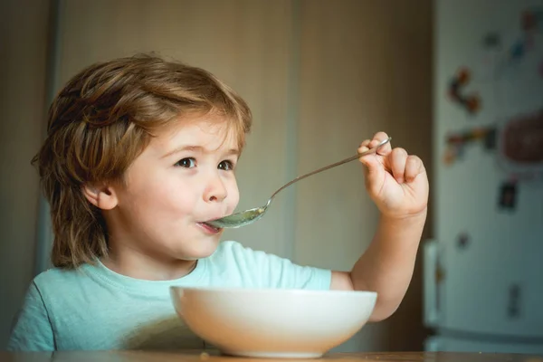 Çocuk yiyor. Mutfakta kahvaltı eden küçük bir çocuk. Ebeveyn -lik. Mutlu çocuk. Genç çocuk yüzünde komik ifade ile yemek masada oturan. — Stok fotoğraf