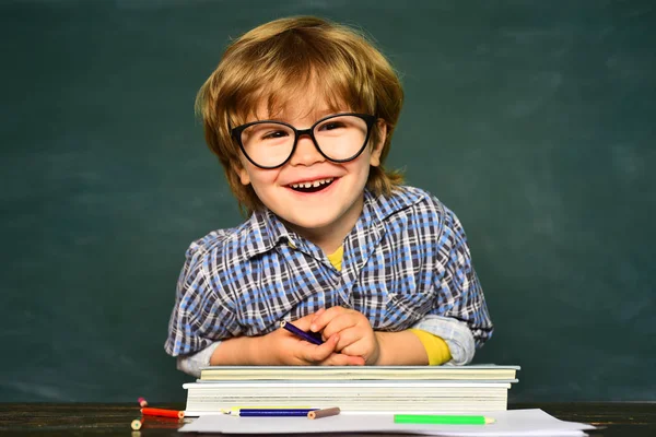 学校に戻って幸せな時間。黒板の背景にメガネをかけた面白い少年。本を持つ子供. — ストック写真
