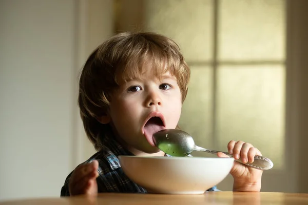 Ein kleines Kind sitzt am Tisch und isst mit lustigem Gesichtsausdruck. Leckeres Kinderfrühstück. Leckeres Kinderfrühstück. Fröhliches Kleinkind isst Lebensmittel selbst mit Löffel. — Stockfoto