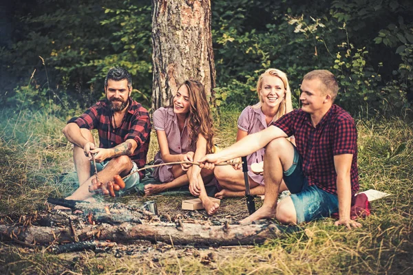 Zwei Paare zelten im Wald. Jungs, die Würstchen über Feuer braten. Lächelnde Freunde am Lagerfeuer am Abend — Stockfoto