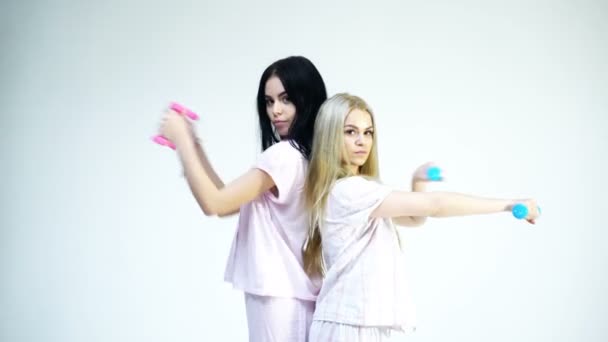 Soeurs ou meilleurs amis en pyjama font du sport. Concept d'exercices matinaux. Les filles tiennent des haltères, isolées sur fond blanc. Blonde et brune sur les visages sérieux se soucient de la figure, faire des exercices . — Video