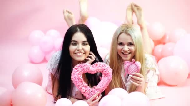 Las niñas yacen cerca de globos, sostiene los juguetes del corazón, fondo rosa. Concepto de San Valentín. Rubia y morena en caras sonrientes soñando con amor y cita. Hermanas, amigas en pijama en pijama . — Vídeo de stock
