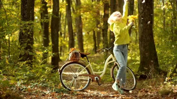 Herbst- und Blattherbstträume. Frau im Herbstkleid mit fallenden Blättern über dem Hintergrund der Natur. Urlaub im Freien. schöne junge Frau auf dem Fahrrad im Park. — Stockvideo