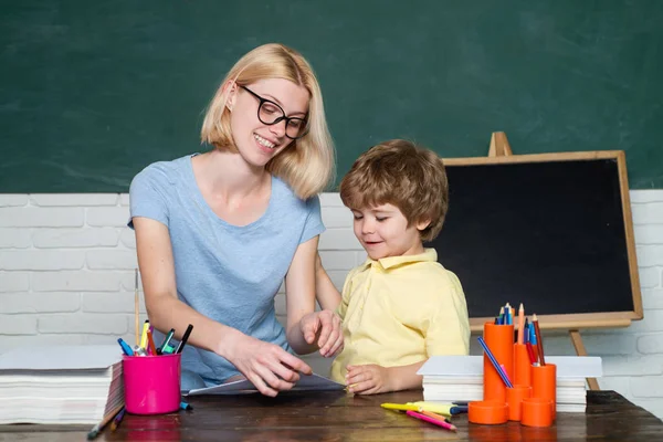 Anne okulda sınıfta oğluna ders veriyor. Komik küçük çocuk ve genç kadın öğretmen kara tahta arka plan üzerinde eğleniyor. Okul çocukları. — Stok fotoğraf