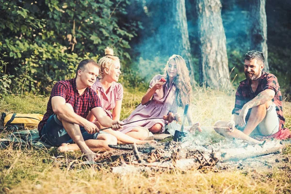 Grupo de amigos acampando na floresta. Dois casais a cozinhar salsichas ao lume. Morena e loira meninas relaxando com seus namorados ao ar livre — Fotografia de Stock