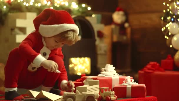 Glückliches nettes Kind in Weihnachtsmütze mit Geschenk haben ein Weihnachtsfest. fröhliches kleines Kind in Winterkleidung denkt an Weihnachtsmann in der Nähe des Weihnachtsbaums. — Stockvideo