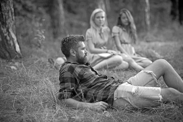 Um tipo barbudo deitado na relva no prado da floresta. Meninas sentadas ao lado da fogueira. Amigos acampar no final do verão. Relaxamento na floresta — Fotografia de Stock