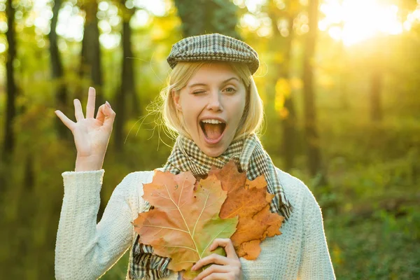 Autumn woman. Happy autumn girl wink. Cheerful carefree autumn woman in park on sunny day. Girl playing with leaves on autumn leaves background. — Stock Photo, Image