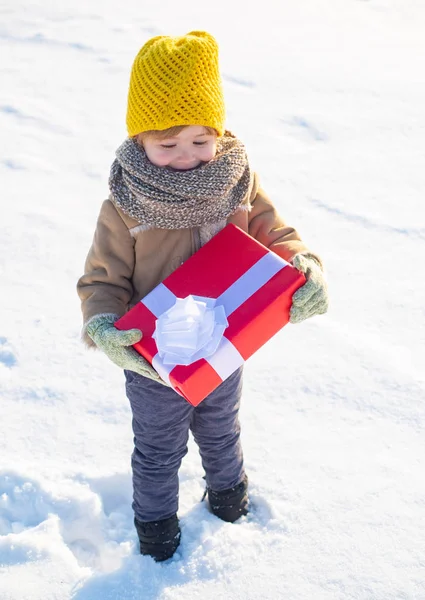 小さな男の子は冬服の帽子とスカーフを間近に着ます。新年おめでとう、メリークリスマス。冬休みのコンセプト。素晴らしい休日をお過ちですか?幸せな冬の子供は贈り物弓雪の背景を保持します — ストック写真