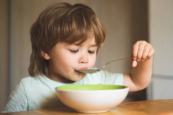 Lachen schattige kind baby jongen zittend in kinderstoel en eten op onscherpe achtergrond. Portret van zoete weinig lachen baby jongen met blonde haren eten van plaat vasthouden lepel-close up. — Stockfoto