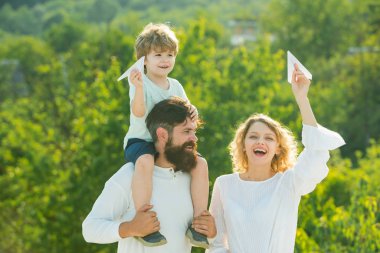Mutlu gülümseyen çocuk omzunda baba kameraya bakıyor. Baba anne ve küçük oğlu dışarıda birlikte oynuyorlar. Mutlu baba omuzlarında omuz aylak yapıyor.