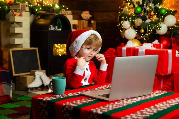 Niño Santa sombrero y traje de divertirse. Comprar regalos de Navidad en línea. Concepto de compras navideñas. Servicio de regalos. Pequeño genio. Santa ayudante. Niño con portátil cerca del árbol de Navidad —  Fotos de Stock