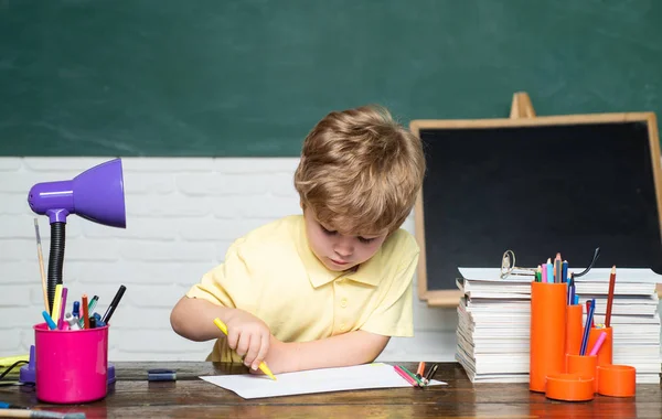 Boldog aranyos szorgalmas gyermek ül egy íróasztal beltéri-iskola fogalmát. Gyermek elemi iskola könyv és táska. Chalkboard másolási hely. — Stock Fotó
