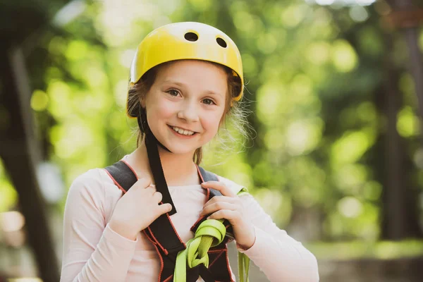 Retrato de uma linda criança em um parque de cordas entre árvores. Menina feliz escalando uma árvore. Capacete - equipamento de segurança para a menina brincando. Parque infantil. Escalada segura esporte extremo com capacete . — Fotografia de Stock