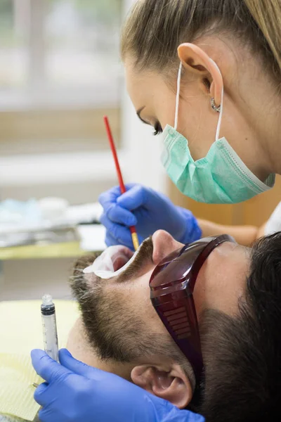 Zbadaj młodego mężczyznę przez dentystę na jasnym, niewyraźne tło. Kobieta dentysta kontroli zębów pacjenta z lustrem w nowoczesnej klinice dentystycznej. Brodaty mężczyzna o zęby badane na dentystów. — Zdjęcie stockowe