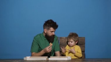 Yeşil gömlekli sakallı adam pizza yiyor. Çocuk pizza yemeyi reddediyor. Adam çocuğu pizza yemeye ikna eder. Abur cubur. Pizza yemek için ayartma. Abur cubur. Mavi arka plan.