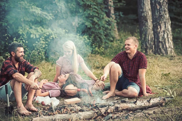 Smiling friends having picnic in woods. Two happy couples enjoying summer evening outdoors, romantic date. Bearded man and his friend frying sausages over fire while their girlfriends rest