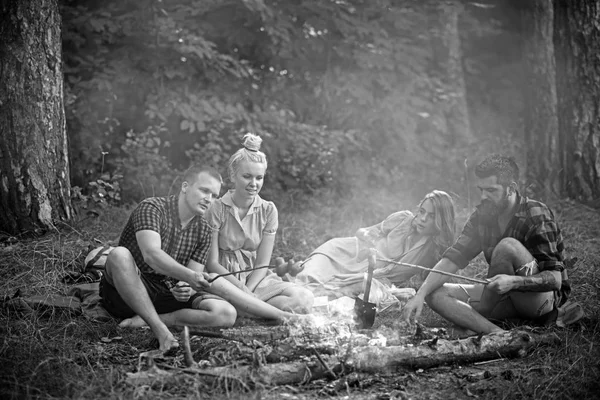 Dois casais a fazer piquenique na floresta. Homem barbudo e seu melhor amigo cozinhando salsichas sobre fogo. Jovens caminhantes ao redor da fogueira à noite. Conceito de amizade e lazer — Fotografia de Stock