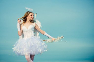 Mavi gökyüzü ve beyaz bulutlara karşı yay ve ok ile ayakta melek kanatları ile Masum Kız. Genç Cupid. Melek kostümü beyaz elbise ve tüy kanatları giyen toddler kız. Aşık küçük melek