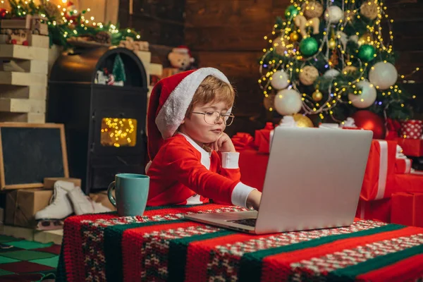 Comprar regalos de Navidad en línea. Concepto de compras navideñas. Servicio de regalos. Pequeño genio. Santa ayudante. Niño con portátil cerca del árbol de Navidad. Pequeño niño santa sombrero y traje de divertirse —  Fotos de Stock