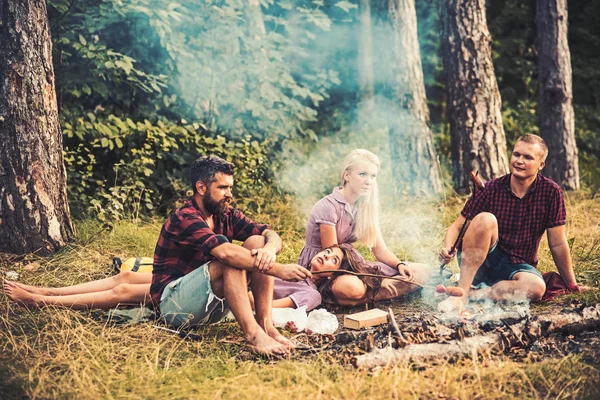 Две пары отдыхают в лесу. Мужчины готовят сосиски на костре. Уставшая брюнетка лежит на коленях блондинок — стоковое фото