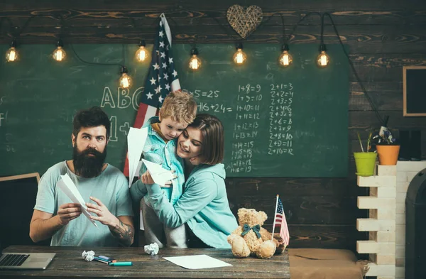 Koncepce knihy. Rodiče učí syna americké tradice hraní. Americká rodina u stolu s synem, takže papír letadla. Dítě s rodiči v učebně s vlajkou usa, tabuli na pozadí — Stock fotografie