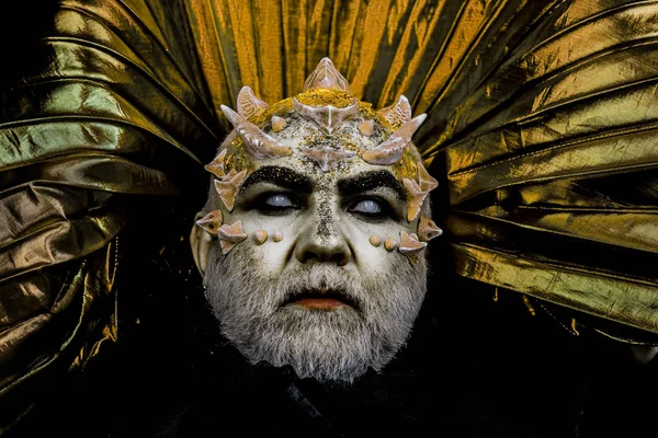 Man with thorns or warts, face covered with glitters. Demon with golden collar on black background. Fantasy concept. Senior man with white beard dressed like monster. Alien, demon, sorcerer makeup.