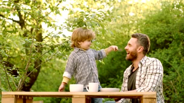 Skratt söt barn pojke sitter i barnstol och äta på grön natur bakgrund. Trädgårdsfest i Amerika. Pappa leker med Kid. Pappa och son leker tillsammans. Pappa och son äter. — Stockvideo