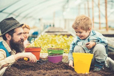 Serada babasıyla oynayan gülümseyen çocuk. Mavi yelekli sakallı adam, sarı gömlek ve şapkalı oğluna bakıyor, anne büyüteçle bitkileri kontrol alıyor. Eko bahçe konsepti