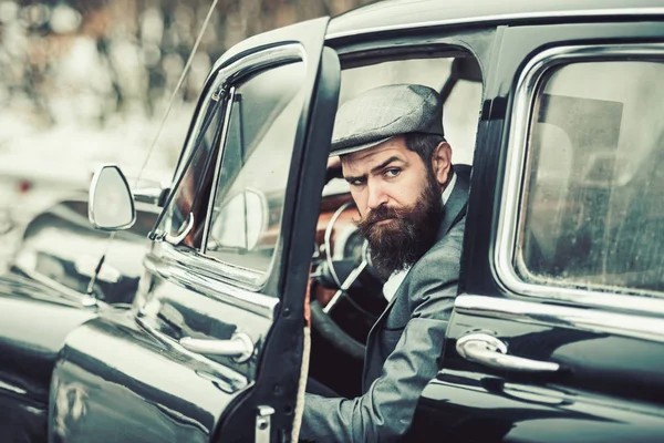 brutal bearded man with a mustache in a cap in the retro car looking at camera.